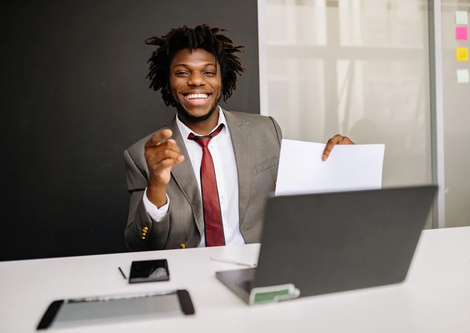 Smiling businessman holding paper at desk with laptop, embodying confidence and professionalism.