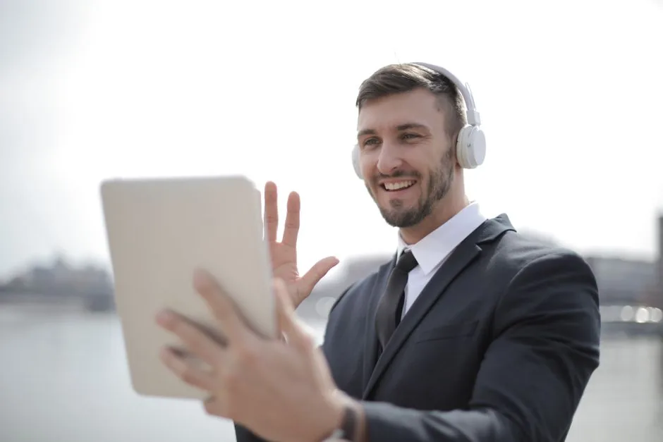 Smiling businessman in suit using a tablet with headphones outdoors, engaging in a video call.